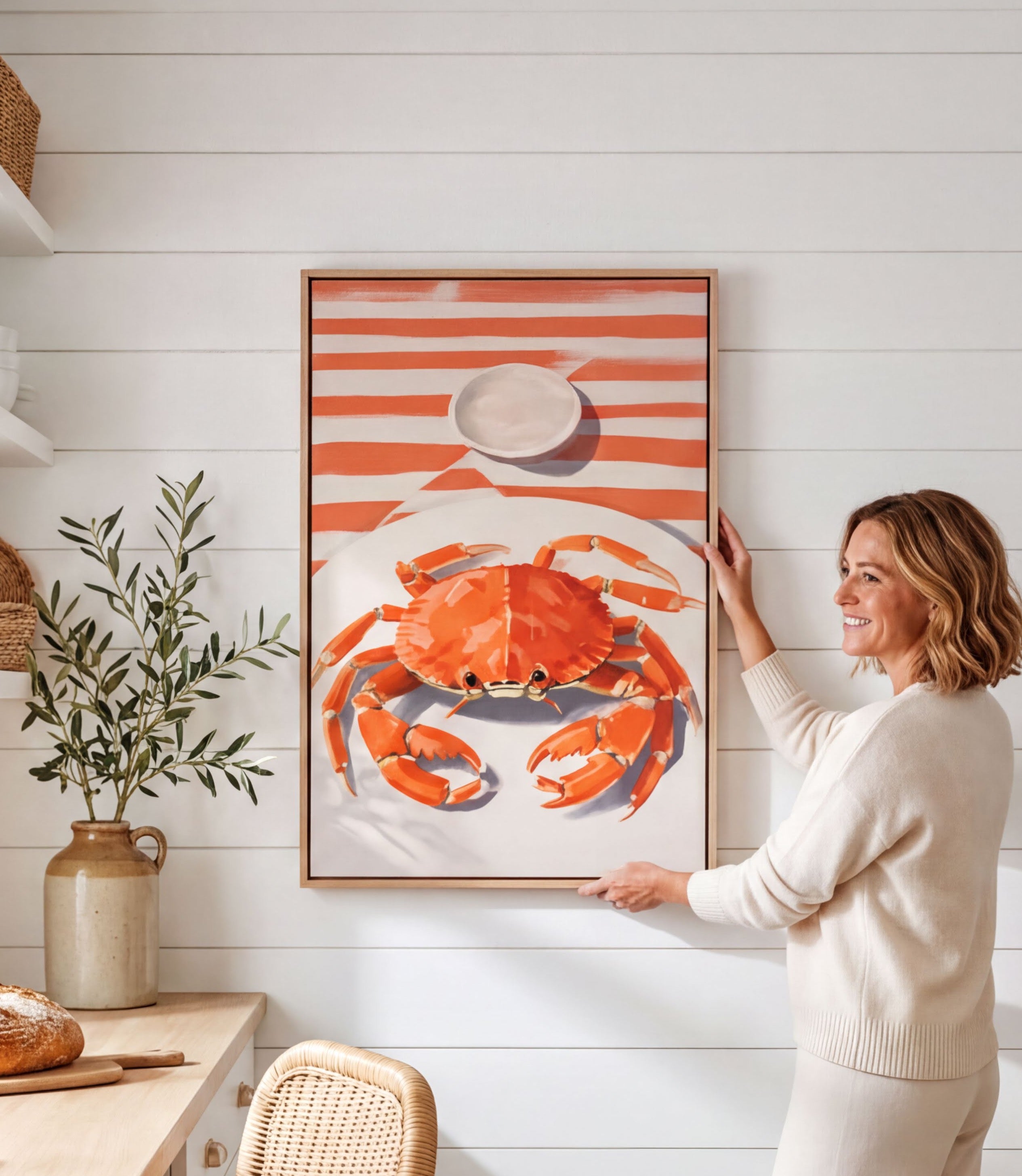 A woman is standing in a kitchen, holding up a framed picture of a crab on a white wall.