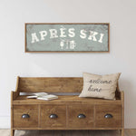 a wooden bench with a "welcome home" pillow on it, set against a white wall. Above the bench, there is a framed sign that says "APRÈS SKI".