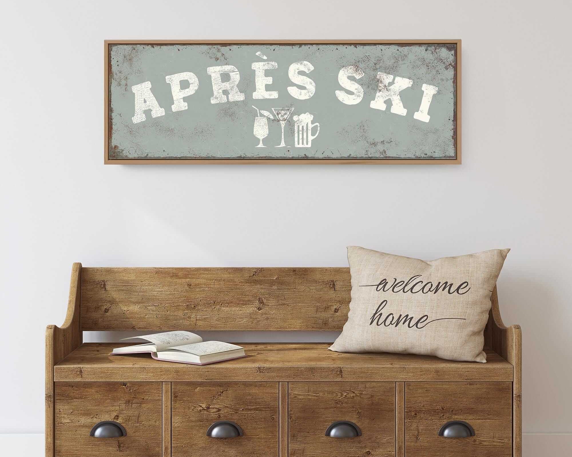 a wooden bench with a "welcome home" pillow on it, set against a white wall. Above the bench, there is a framed sign that says "APRÈS SKI".