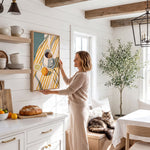 A woman in a beige sweater stands in a cozy kitchen, reaching up to hang a framed picture on the wall. The kitchen features white cabinets, a wooden dining table, and a cat resting on a bench.