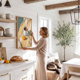 A woman in a beige sweater stands in a cozy kitchen, reaching up to hang a framed picture on the wall. The kitchen features white cabinets, a wooden dining table, and a cat resting on a bench.