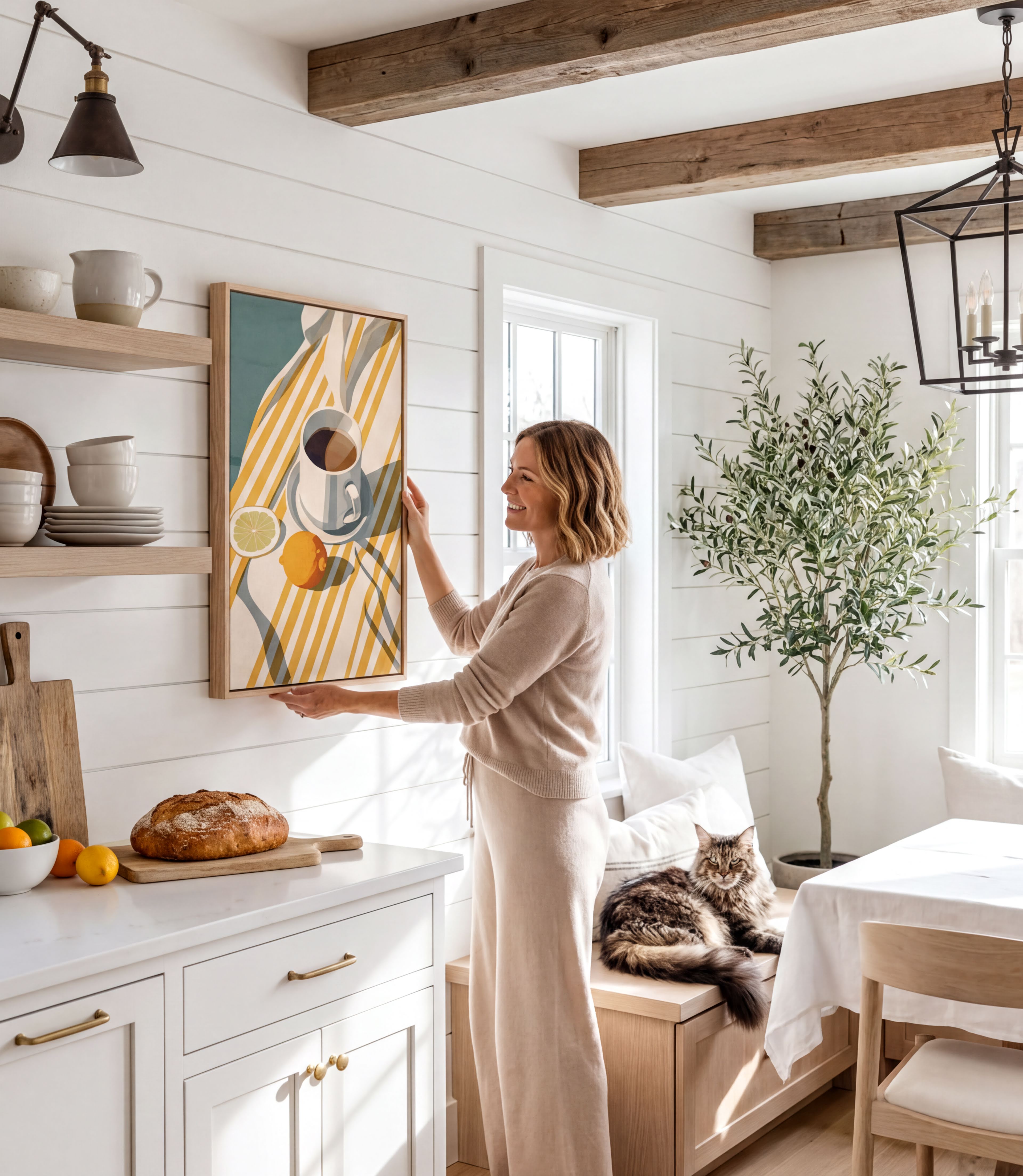 A woman in a beige sweater stands in a cozy kitchen, reaching up to hang a framed picture on the wall. The kitchen features white cabinets, a wooden dining table, and a cat resting on a bench.