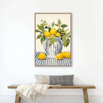 a framed painting of a vase with lemons and green leaves, hanging on a white wall above a wooden bench with a gray pillow.