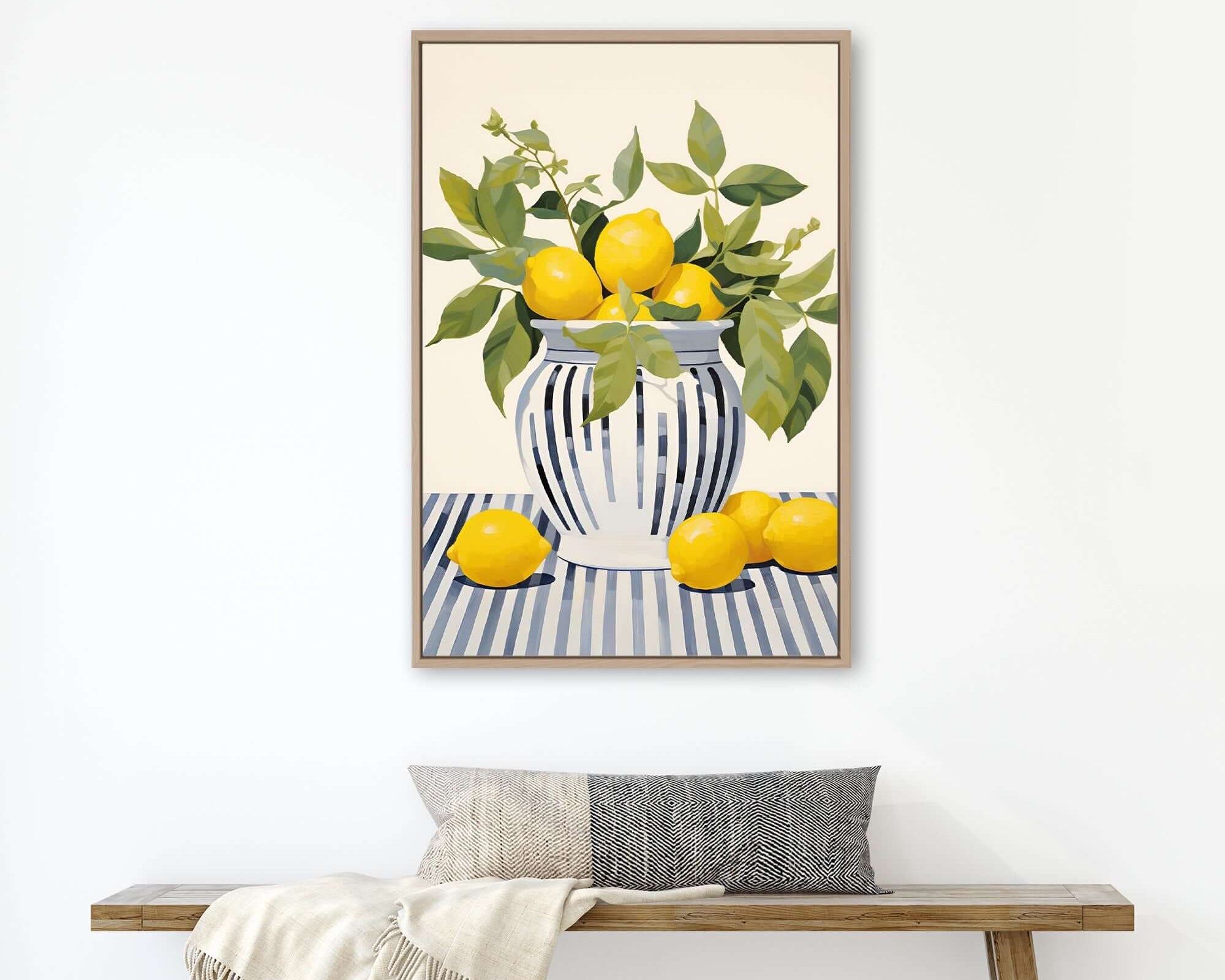 a framed painting of a vase with lemons and green leaves, hanging on a white wall above a wooden bench with a gray pillow.