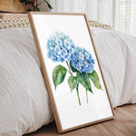 A framed watercolor painting of blue hydrangea flowers is displayed on a wooden floor in a bedroom setting.