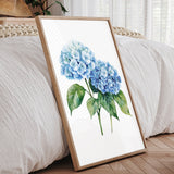 A framed watercolor painting of blue hydrangea flowers is displayed on a wooden floor in a bedroom setting.