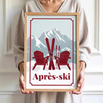 A person is holding a framed print with a winter scene featuring two red Adirondack chairs and skis, with a mountain backdrop.