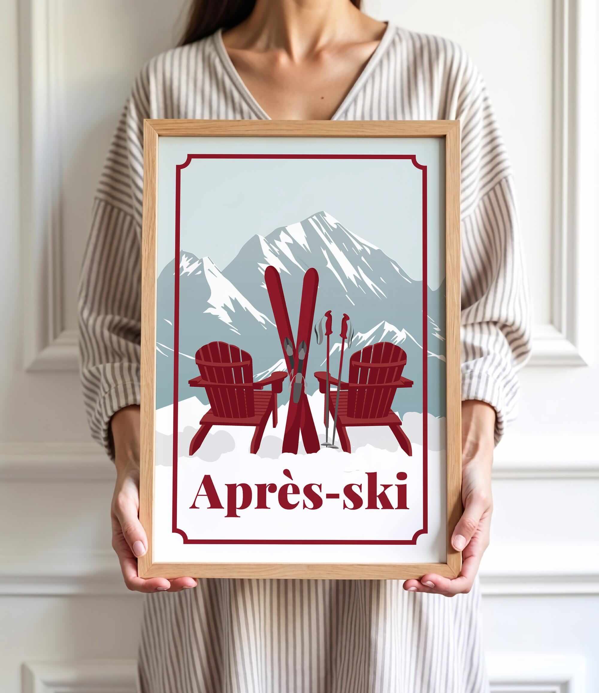 A person is holding a framed print with a winter scene featuring two red Adirondack chairs and skis, with a mountain backdrop.