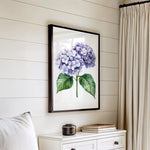 a framed painting of purple hydrangeas hanging on a white wall above a white dresser with a stack of books on top.