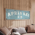 a cozy living room with a wooden wall and a framed sign that reads "APRÈS SKI". The sign is hanging above a couch with pillows, creating a warm and inviting atmosphere.