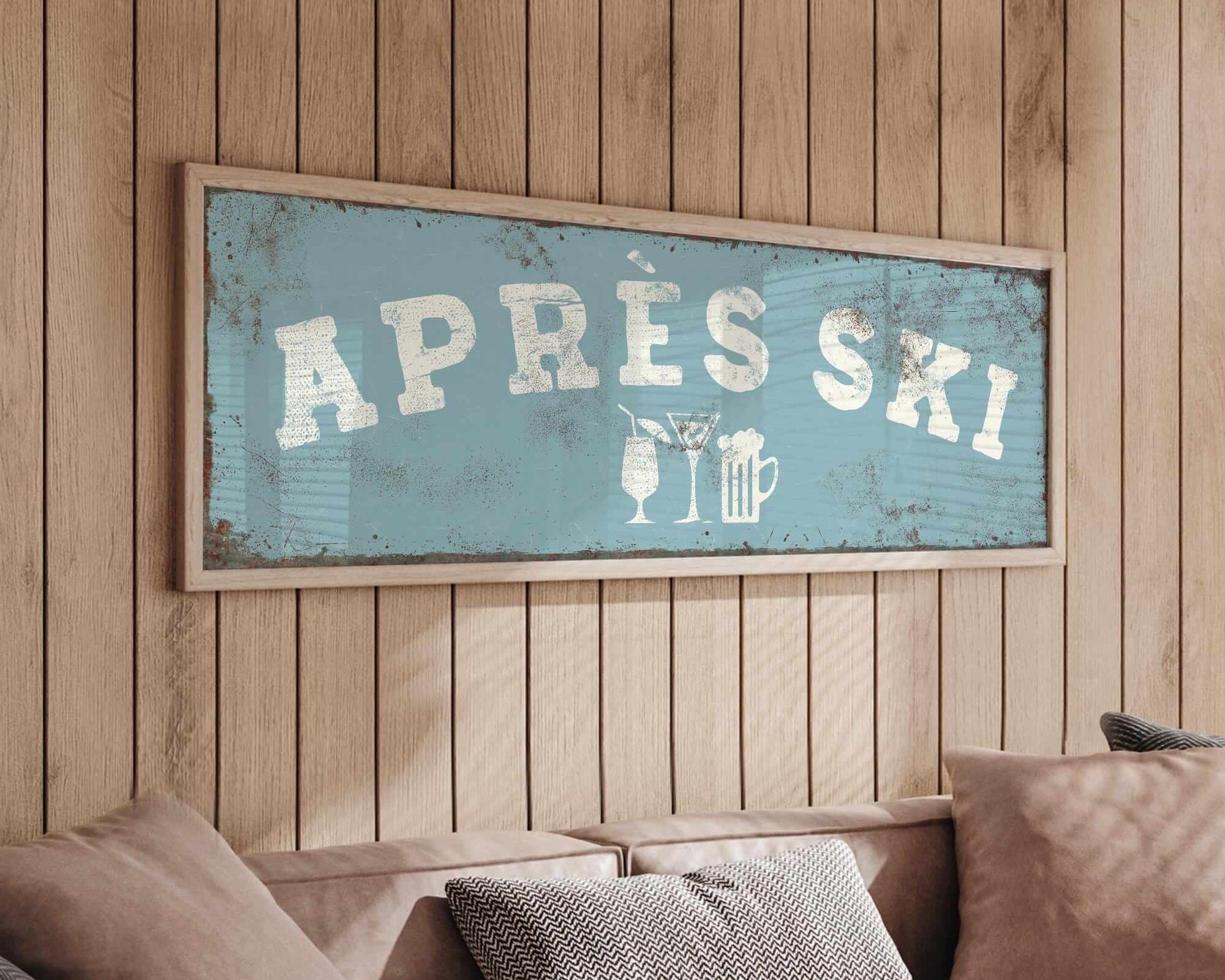 a cozy living room with a wooden wall and a framed sign that reads "APRÈS SKI". The sign is hanging above a couch with pillows, creating a warm and inviting atmosphere.