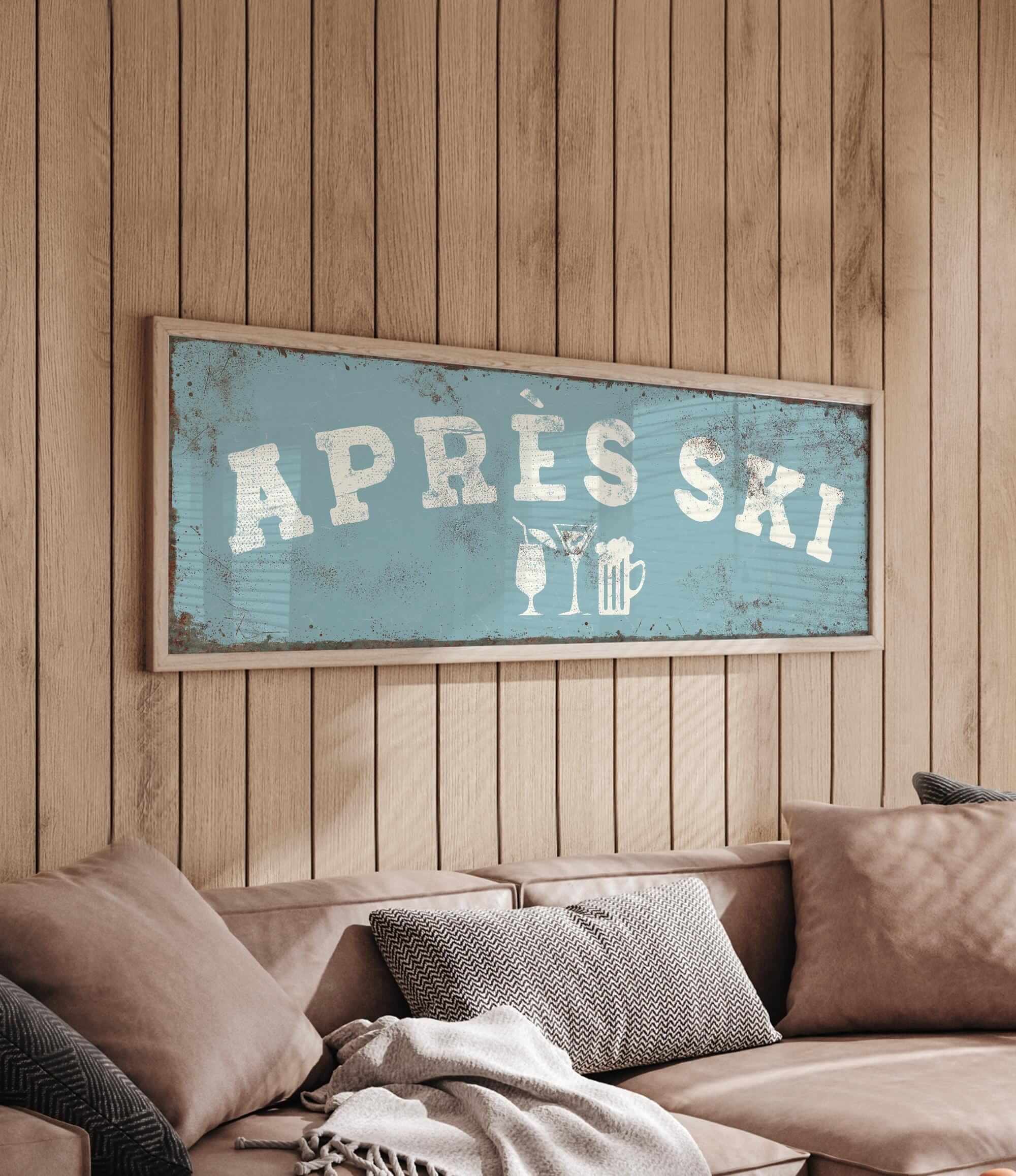 a cozy living room with a wooden wall and a framed sign that reads "APRÈS SKI". The sign is hanging above a couch with pillows, creating a warm and inviting atmosphere.