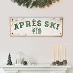 a white fireplace mantel decorated with various holiday items, including a sign that says "APRES SKI", candles, pine cones, and a glass dome.