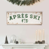 a white fireplace mantel decorated with various holiday items, including a sign that says "APRES SKI", candles, pine cones, and a glass dome.