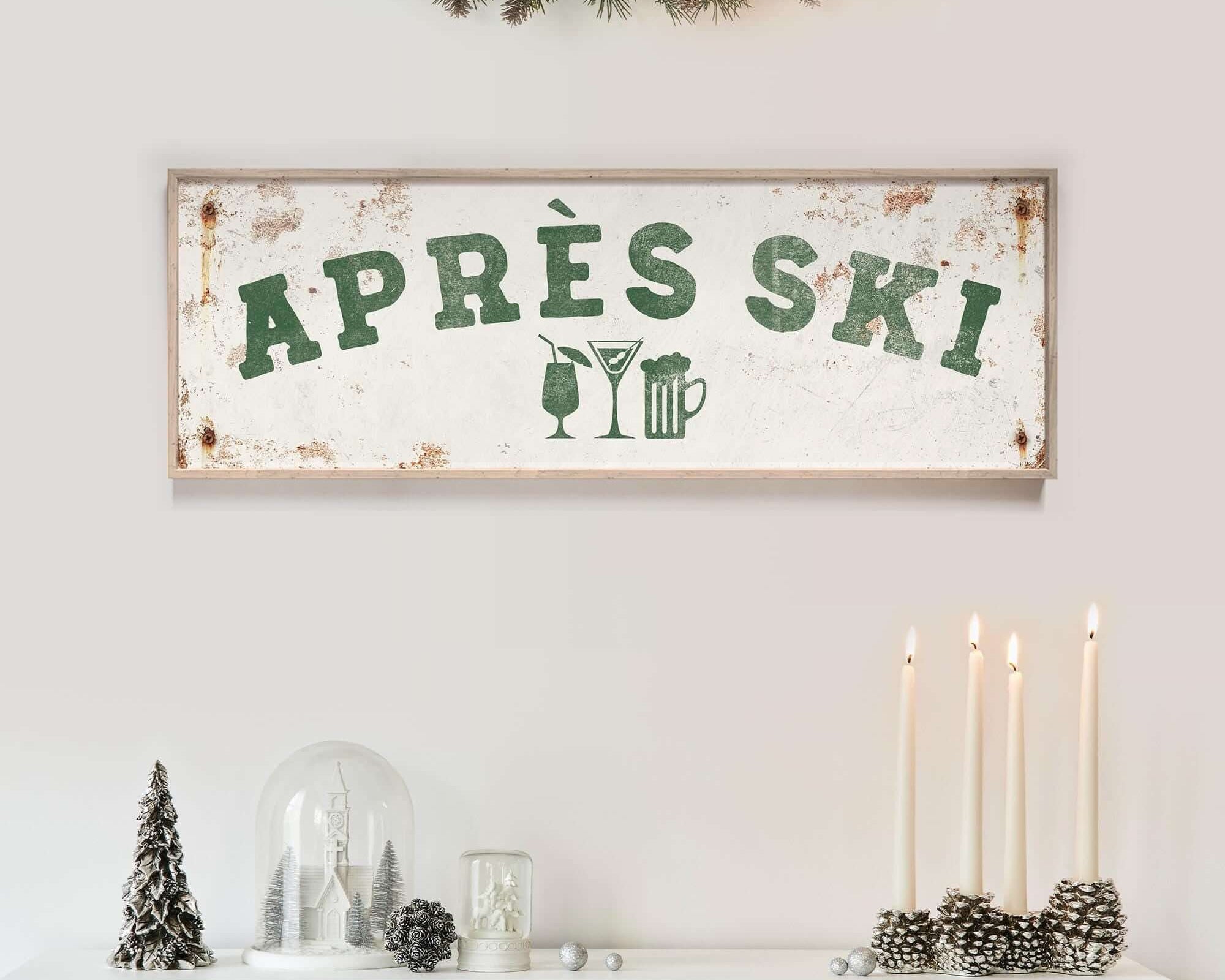 a white fireplace mantel decorated with various holiday items, including a sign that says "APRES SKI", candles, pine cones, and a glass dome.