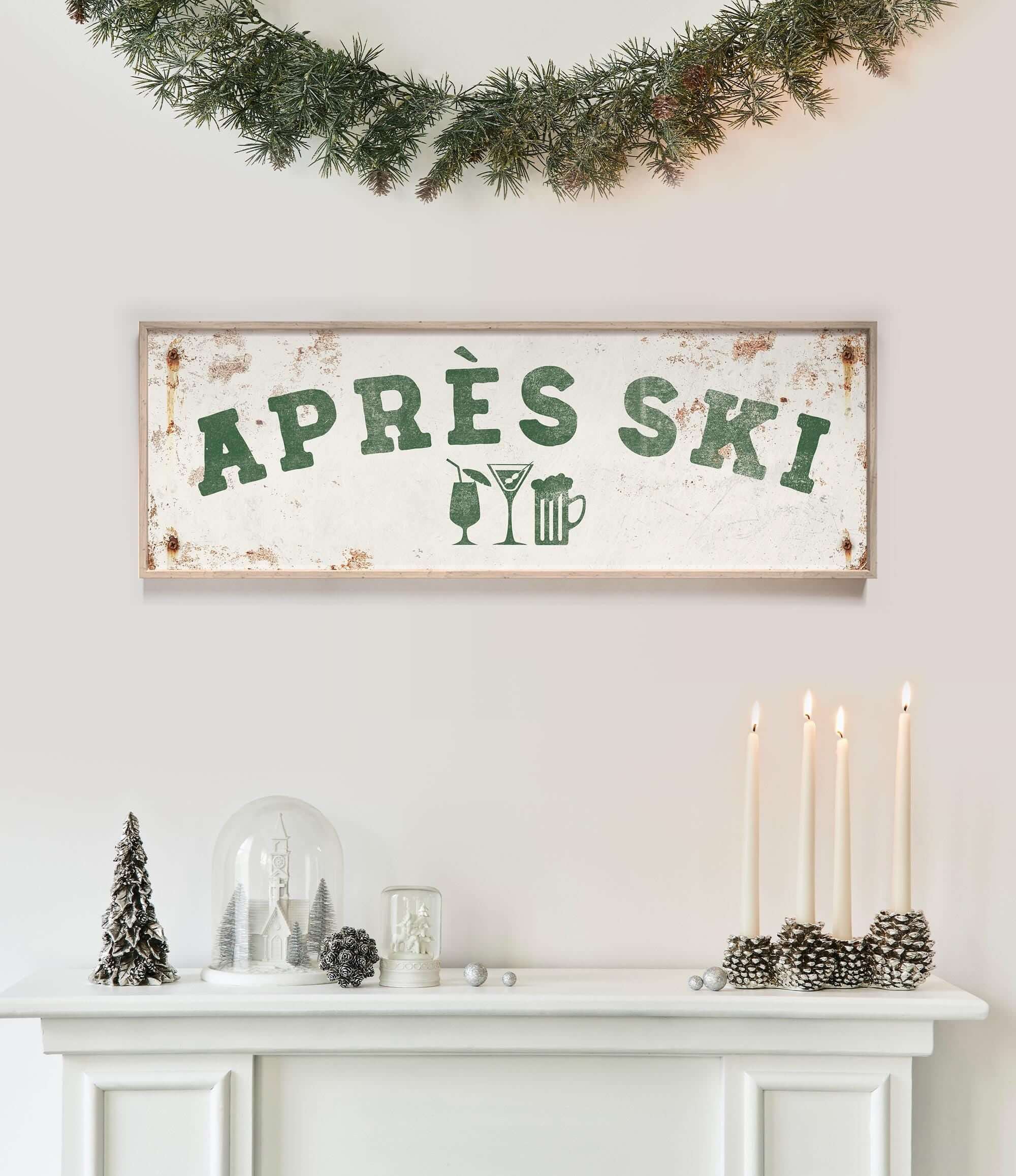a white fireplace mantel decorated with various holiday items, including a sign that says "APRES SKI", candles, pine cones, and a glass dome.