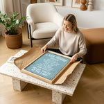 A woman is sitting on the floor in a living room, holding a framed sign with text on it.