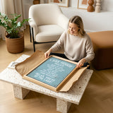 A woman is sitting on the floor in a living room, holding a framed sign with text on it.