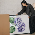 A woman is standing next to a large framed painting of purple hydrangeas, examining the artwork.
