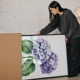 A woman is standing next to a large framed painting of purple hydrangeas, examining the artwork.