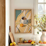 a cozy kitchen scene with a framed painting of a cup of coffee and a lemon on a striped tablecloth, surrounded by various kitchen items such as a cutting board, a bowl of fruit, and a vase of flowers.
