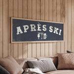 a cozy living room with a wooden wall and a comfortable couch. Above the couch, there is a framed sign that reads "APRÈS SKI", which appears to be a humorous take on skiing.