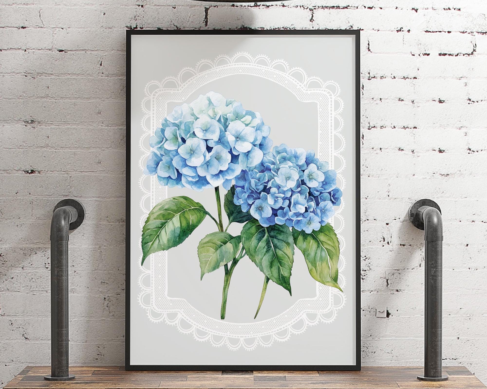 A framed watercolor painting of blue hydrangeas is displayed on a wooden shelf against a white brick wall, with a metal light fixture above it.