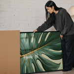 A woman in a gray sweater is leaning over a large framed print of a large green leaf with gold accents, examining it closely.