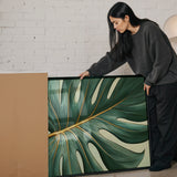 A woman in a gray sweater is leaning over a large framed print of a large green leaf with gold accents, examining it closely.