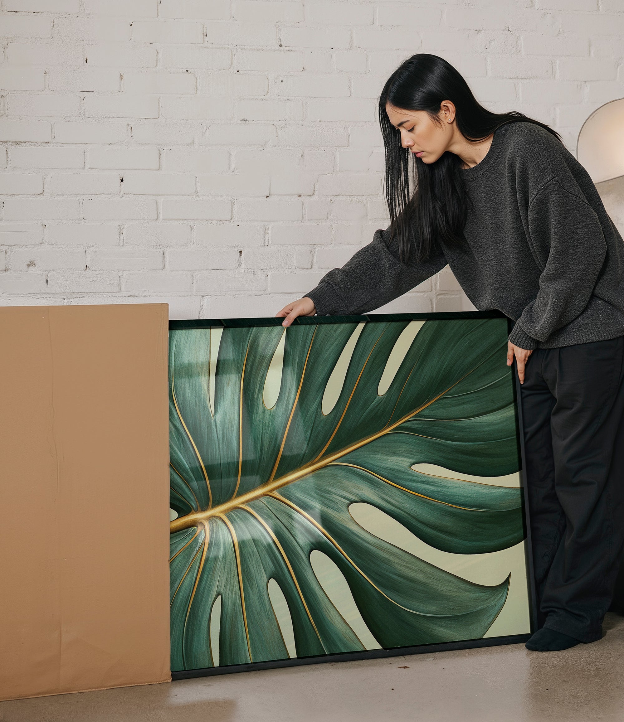 A woman in a gray sweater is leaning over a large framed print of a large green leaf with gold accents, examining it closely.