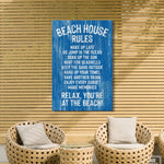 Two wicker chairs and a small table sit on a patio with beach decor, including a charming "Beach House Rules Sign, Painted Wood, Ocean Blue" displayed on a wooden slat wall.