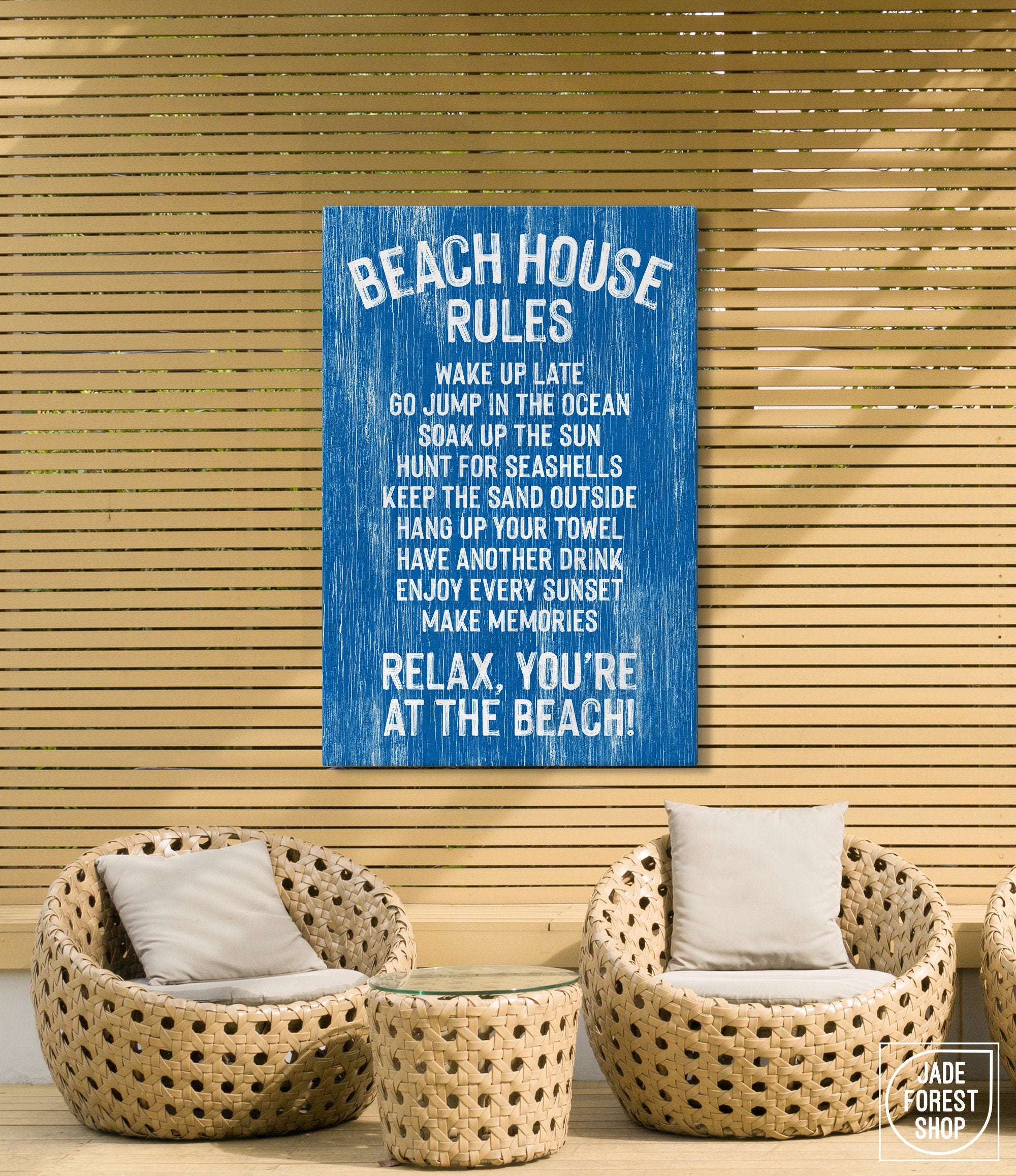 Two wicker chairs and a small table sit on a patio with beach decor, including a charming "Beach House Rules Sign, Painted Wood, Ocean Blue" displayed on a wooden slat wall.