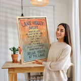 A woman is holding up a framed sign with a list of rules for a lake house, smiling and posing for the camera.