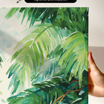 a close-up view of a vibrant, lush tropical plant with large, green leaves. The plant appears to be a type of palm or fern, with intricate patterns and textures. The background is a light green color, providing a contrast to the vivid foliage. The image is described as an "unframed canvas print" and "a hand holding it", suggesting that it is a printed artwork being held by someone.