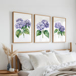 a bedroom with a bed, white pillows, and a wooden headboard. Above the bed, there are three framed watercolor paintings of purple hydrangeas hanging on the wall.