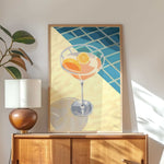 a framed illustration of a cocktail glass with orange slices, placed on a wooden cabinet or dresser.