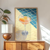 a framed illustration of a cocktail glass with orange slices, placed on a wooden cabinet or dresser.