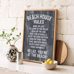 a chalkboard sign with beach house rules, accompanied by a wooden board, a vase with a plant, and a bowl of lemons, all placed on a white countertop.