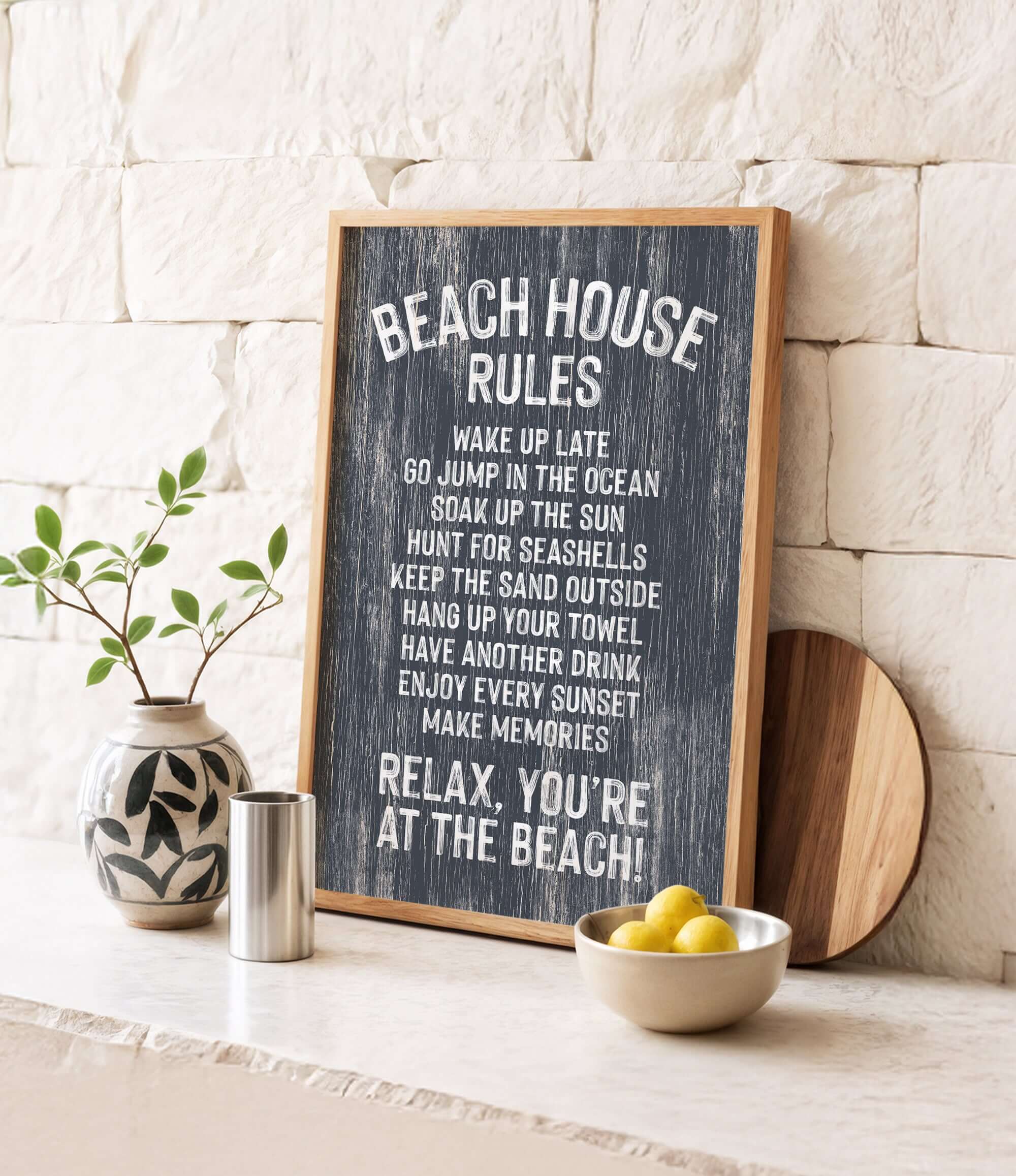 a chalkboard sign with beach house rules, accompanied by a wooden board, a vase with a plant, and a bowl of lemons, all placed on a white countertop.