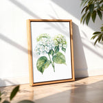 A framed watercolor painting of white hydrangea flowers is displayed on a wooden floor against a white wall, with a plant visible in the background.