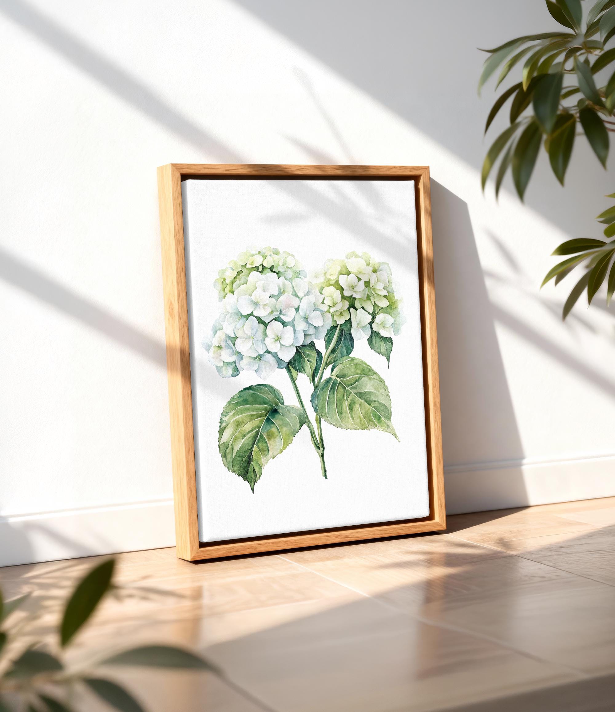 A framed watercolor painting of white hydrangea flowers is displayed on a wooden floor against a white wall, with a plant visible in the background.