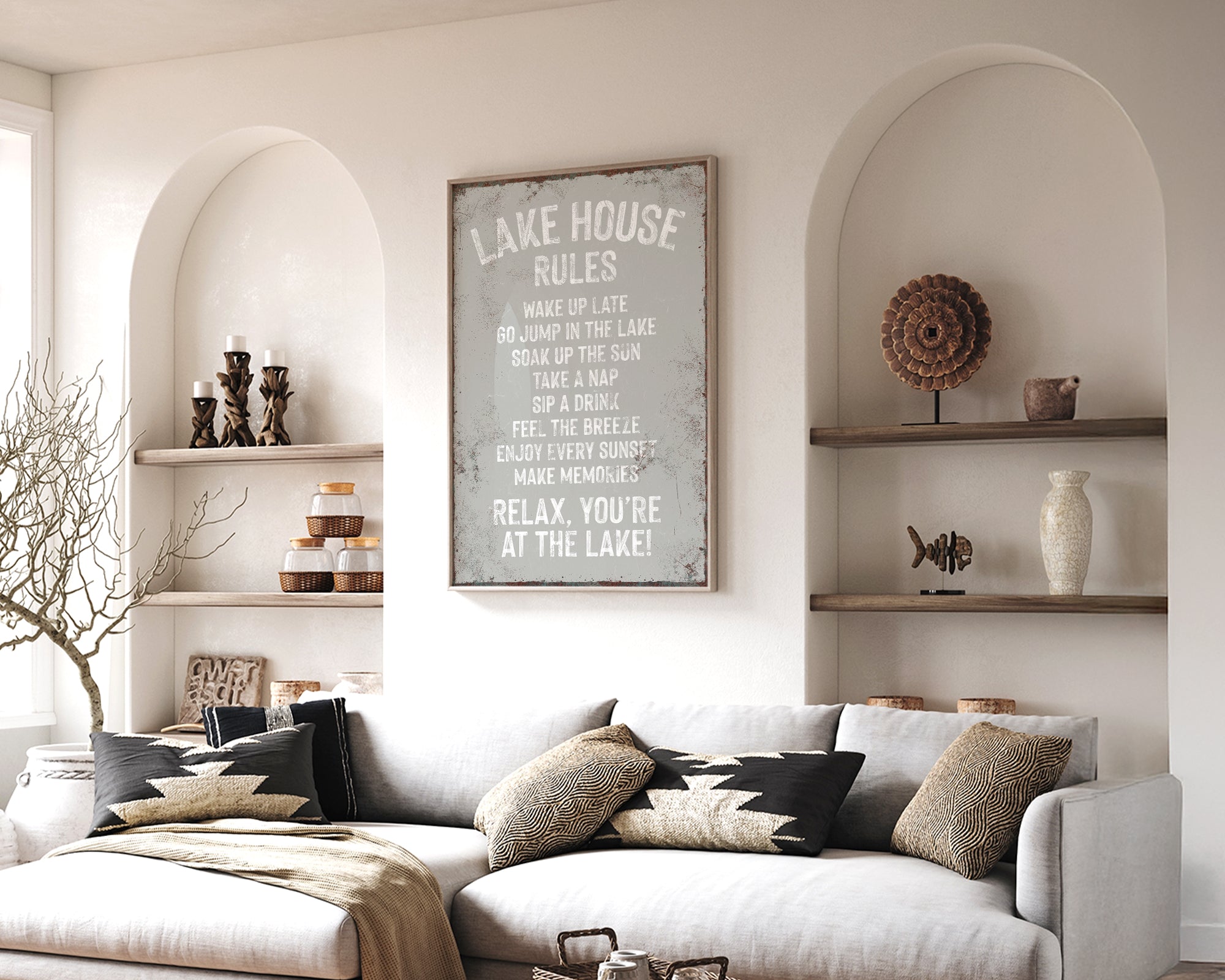 a cozy living room with a white sofa, a wooden coffee table, and a framed sign that reads "LAKESHORE RULES" above the sofa. The room has arched doorways, and there are shelves displaying decorative items and a vase on the wall.