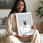 A woman in a beige outfit is sitting on a bed and holding up a framed poster with a dog in a bathtub.