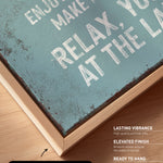 a close-up view of a textured, weathered canvas print with a wooden frame. The text on the canvas is in white and reads "Enjoy life, make memories, relax at the lake".