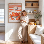 A woman in a beige outfit is leaning against a wall, looking at her phone in a bright and airy kitchen with a large painting of a crab on the wall.
