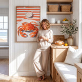 A woman in a beige outfit is leaning against a wall, looking at her phone in a bright and airy kitchen with a large painting of a crab on the wall.