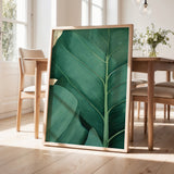 a large framed print of a green leaf with intricate patterns, displayed on a wooden floor in a room with a dining table and chairs.