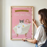 A woman is hanging a framed poster on a wall, which features a cat sitting in a bathtub filled with bubbles.