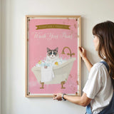 A woman is hanging a framed poster on a wall, which features a cat sitting in a bathtub filled with bubbles.