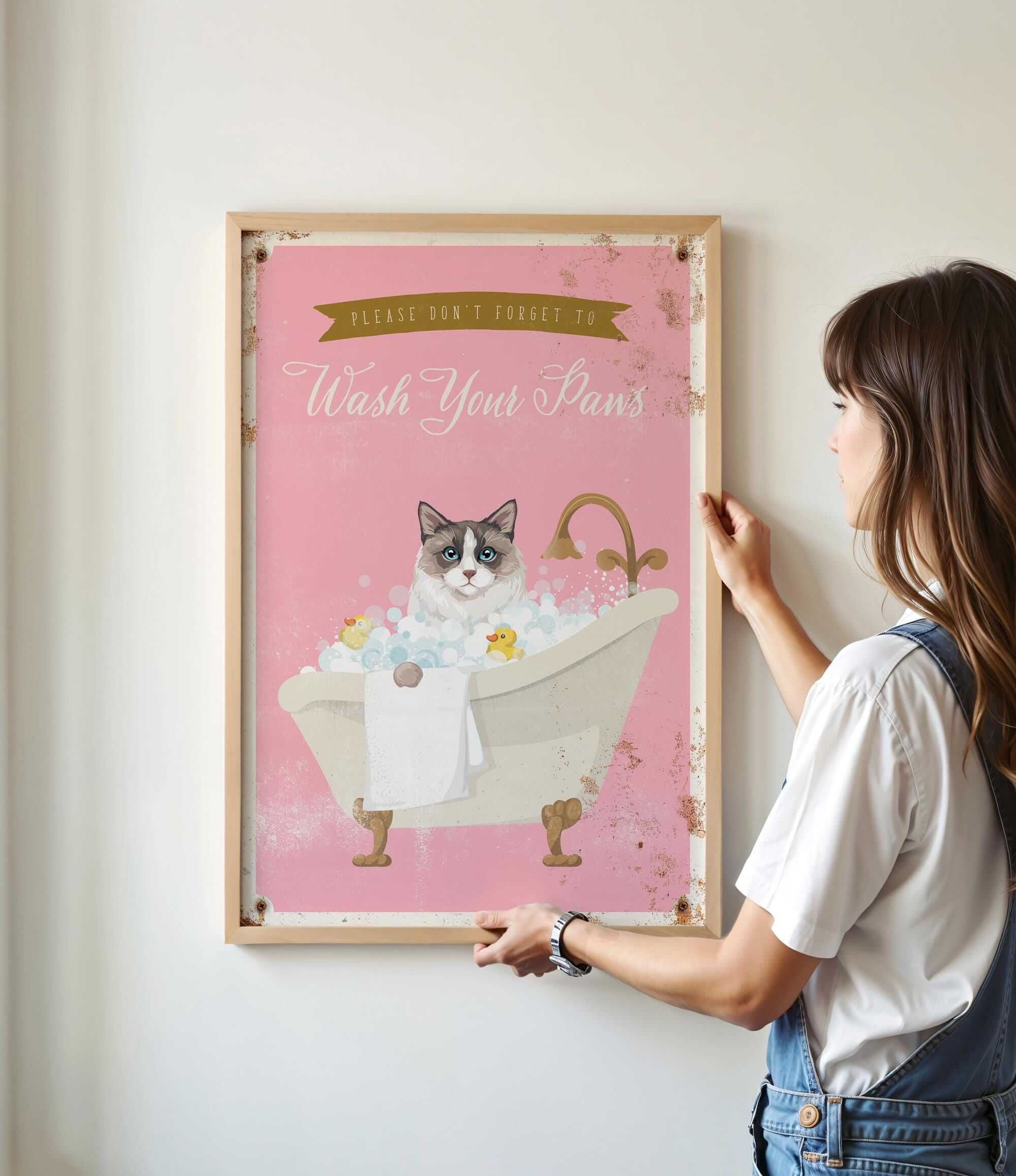 A woman is hanging a framed poster on a wall, which features a cat sitting in a bathtub filled with bubbles.